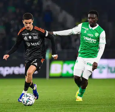 Soccer players contest the ball on a green field; one in black with orange accents, the other in green and white.