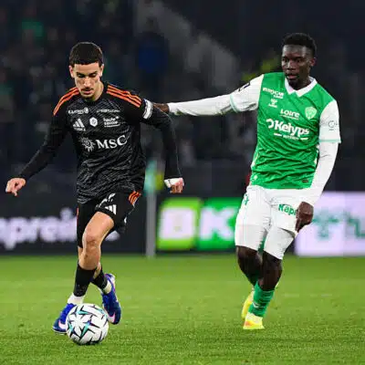 Soccer players contest the ball on a green field; one in black with orange accents, the other in green and white.
