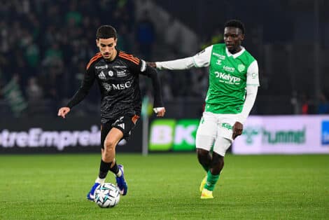 Soccer players contest the ball on a green field; one in black with orange accents, the other in green and white.