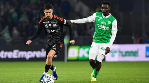 Soccer players contest the ball on a green field; one in black with orange accents, the other in green and white.