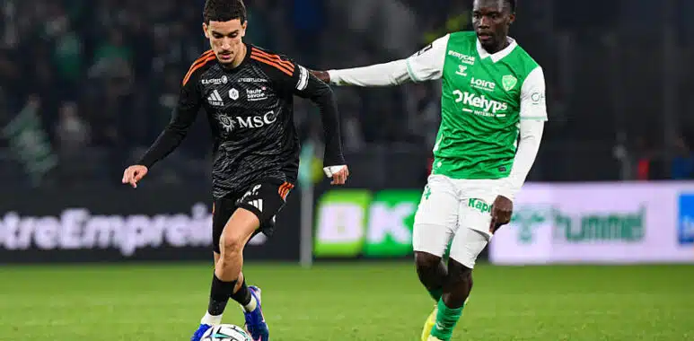 Soccer players contest the ball on a green field; one in black with orange accents, the other in green and white.
