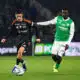 Soccer players contest the ball on a green field; one in black with orange accents, the other in green and white.