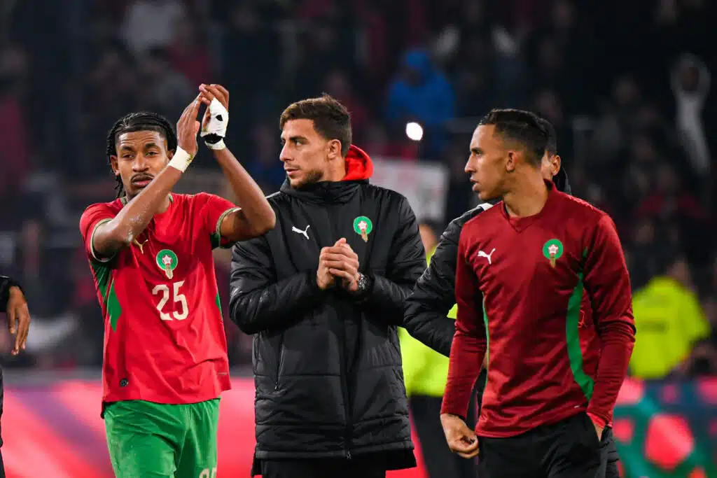 Three Moroccan footballers celebrate on the pitch, red and green uniforms, high-fiving amid a cheering crowd.