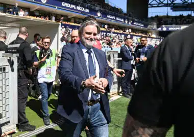 Man in a navy suit and jeans walks toward the camera at a crowded sports stadium, security barriers and photographers nearby.
