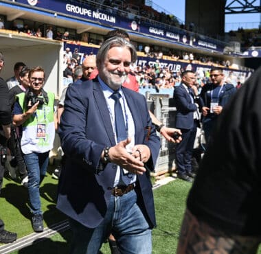 Man in a navy suit and jeans walks toward the camera at a crowded sports stadium, security barriers and photographers nearby.
