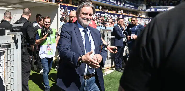 Man in a navy suit and jeans walks toward the camera at a crowded sports stadium, security barriers and photographers nearby.