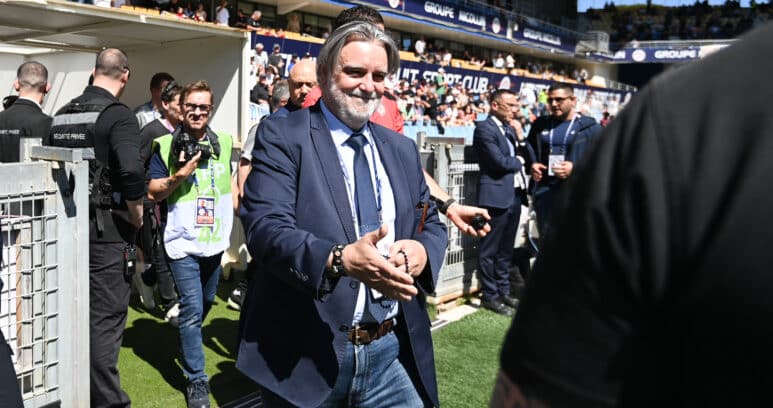 Man in a navy suit and jeans walks toward the camera at a crowded sports stadium, security barriers and photographers nearby.