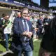 Man in a navy suit and jeans walks toward the camera at a crowded sports stadium, security barriers and photographers nearby.