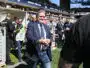 Man in a navy suit and jeans walks toward the camera at a crowded sports stadium, security barriers and photographers nearby.