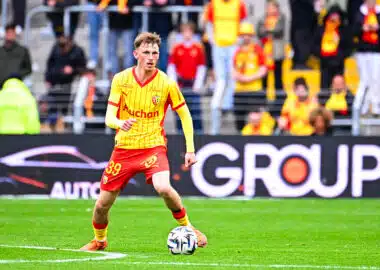 Soccer player in a yellow jersey and red shorts dribbles a ball on a green pitch with spectators in the stands behind him.