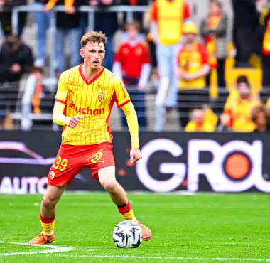 Soccer player in a yellow jersey and red shorts dribbles a ball on a green pitch with spectators in the stands behind him.