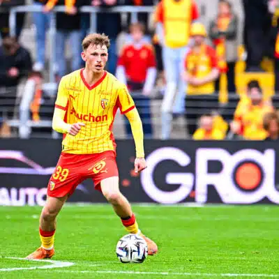 Soccer player in a yellow jersey and red shorts dribbles a ball on a green pitch with spectators in the stands behind him.