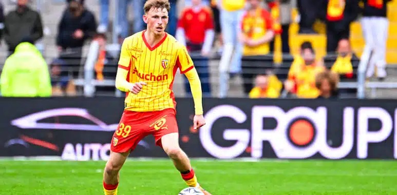 Oscar Lenne | AllezPaillade.com Soccer player in a yellow jersey and red shorts dribbles a ball on a green pitch with spectators in the stands behind him.