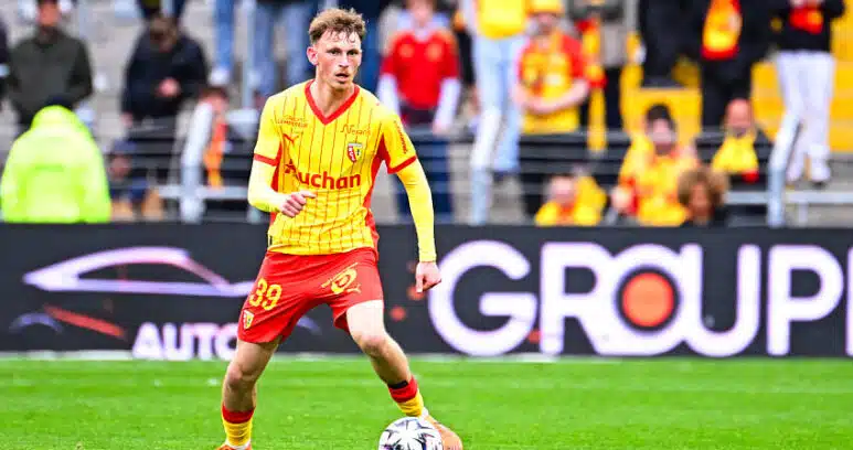 Soccer player in a yellow jersey and red shorts dribbles a ball on a green pitch with spectators in the stands behind him.