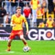Soccer player in a yellow jersey and red shorts dribbles a ball on a green pitch with spectators in the stands behind him.