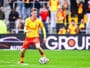 Soccer player in a yellow jersey and red shorts dribbles a ball on a green pitch with spectators in the stands behind him.