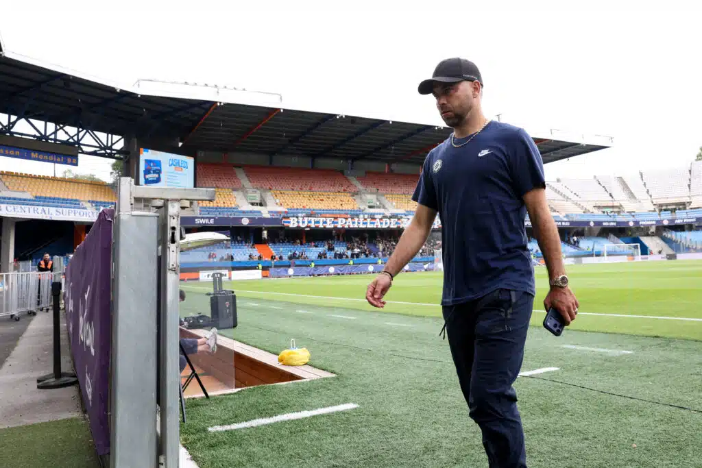 Man in a navy Nike shirt and cap walking along the stadium sideline, holding a smartphone.