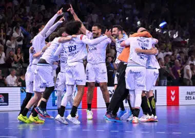 A group of white-uniform handball players celebrate together in a circle on a blue court, crowd cheering in the stands behind them.