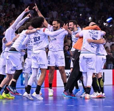 A group of white-uniform handball players celebrate together in a circle on a blue court, crowd cheering in the stands behind them.