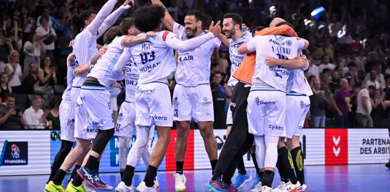 Montpellier Handball v HBC Nantes - Finale Coupe de France National Masculine 202526 | AllezPaillade.com A group of white-uniform handball players celebrate together in a circle on a blue court, crowd cheering in the stands behind them.