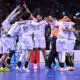 A group of white-uniform handball players celebrate together in a circle on a blue court, crowd cheering in the stands behind them.