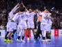 A group of white-uniform handball players celebrate together in a circle on a blue court, crowd cheering in the stands behind them.