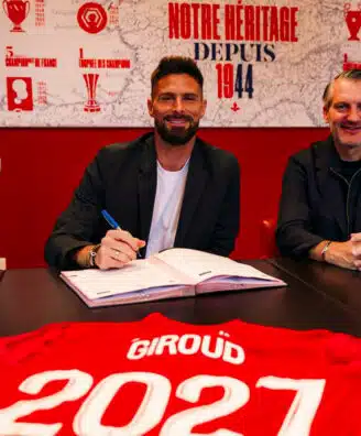 Two men sit at a dark table in a red room, one signing papers while the other smiles; a red sports jersey with &lsquo;GIR0UD&rsquo; and &lsquo;2021&rsquo; is laid in the foreground.