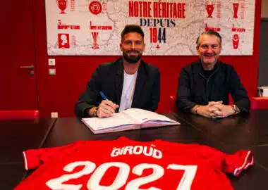 Two men sit at a dark table in a red room, one signing papers while the other smiles; a red sports jersey with &lsquo;GIR0UD&rsquo; and &lsquo;2021&rsquo; is laid in the foreground.