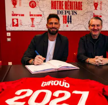 Two men sit at a dark table in a red room, one signing papers while the other smiles; a red sports jersey with &lsquo;GIR0UD&rsquo; and &lsquo;2021&rsquo; is laid in the foreground.