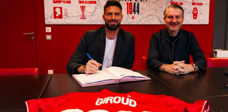 LOSC Lille - Olivier Giroud - Contract extension 08062026 | AllezPaillade.com Two men sit at a dark table in a red room, one signing papers while the other smiles; a red sports jersey with ‘GIR0UD’ and ‘2021’ is laid in the foreground.