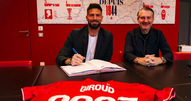 Two men sit at a dark table in a red room, one signing papers while the other smiles; a red sports jersey with &lsquo;GIR0UD&rsquo; and &lsquo;2021&rsquo; is laid in the foreground.