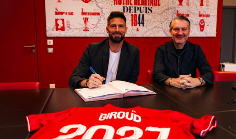 Two men sit at a dark table in a red room, one signing papers while the other smiles; a red sports jersey with &lsquo;GIR0UD&rsquo; and &lsquo;2021&rsquo; is laid in the foreground.