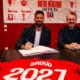 Two men sit at a dark table in a red room, one signing papers while the other smiles; a red sports jersey with &lsquo;GIR0UD&rsquo; and &lsquo;2021&rsquo; is laid in the foreground.