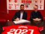 Two men sit at a dark table in a red room, one signing papers while the other smiles; a red sports jersey with &lsquo;GIR0UD&rsquo; and &lsquo;2021&rsquo; is laid in the foreground.