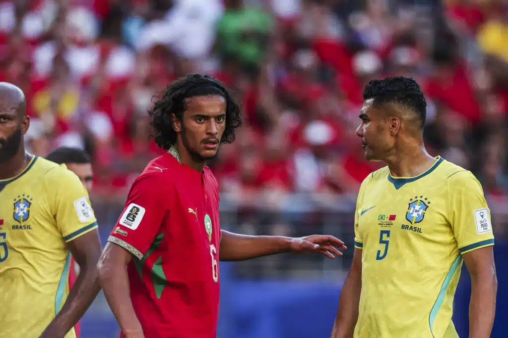 Two Brazil players face off on the field; one in red and one in yellow, with a crowded stadium in the background.