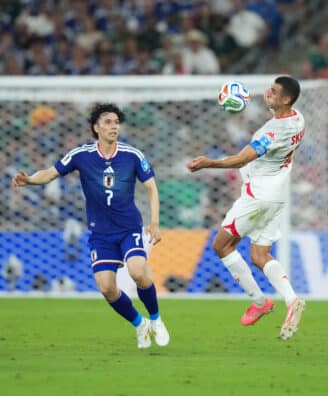 Soccer match action: a white-jersey player jumps to control the ball as blue-jersey defenders approach near the goal in a crowded stadium.