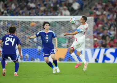 Soccer match action: a white-jersey player jumps to control the ball as blue-jersey defenders approach near the goal in a crowded stadium.