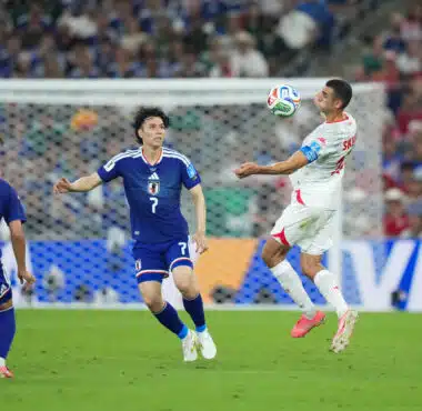 Soccer match action: a white-jersey player jumps to control the ball as blue-jersey defenders approach near the goal in a crowded stadium.