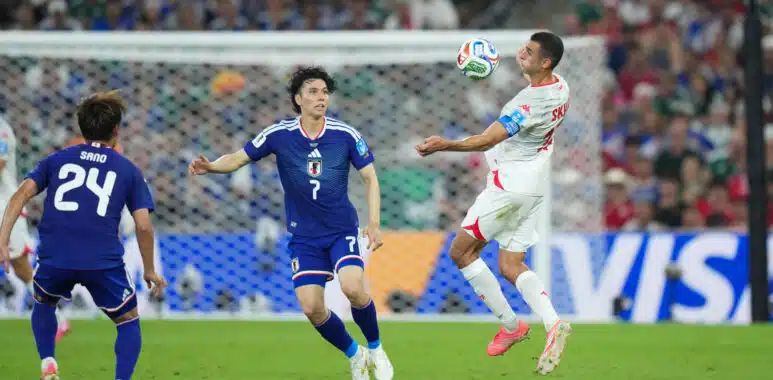 Soccer match action: a white-jersey player jumps to control the ball as blue-jersey defenders approach near the goal in a crowded stadium.