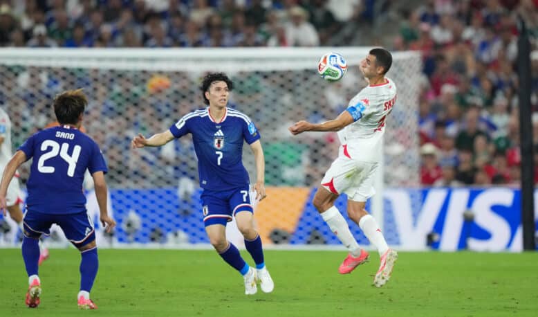 Soccer match action: a white-jersey player jumps to control the ball as blue-jersey defenders approach near the goal in a crowded stadium.