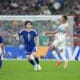 Soccer match action: a white-jersey player jumps to control the ball as blue-jersey defenders approach near the goal in a crowded stadium.