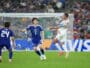 Soccer match action: a white-jersey player jumps to control the ball as blue-jersey defenders approach near the goal in a crowded stadium.