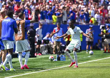 Soccer player in light kit celebrates near the touchline as teammates applaud and fans cheer in the background during a match under sunny skies.