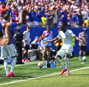 Soccer player in light kit celebrates near the touchline as teammates applaud and fans cheer in the background during a match under sunny skies.