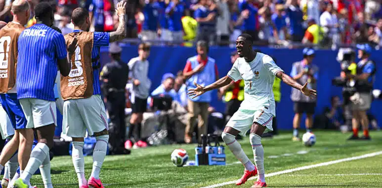 Soccer player in light kit celebrates near the touchline as teammates applaud and fans cheer in the background during a match under sunny skies.