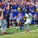 Soccer player in light kit celebrates near the touchline as teammates applaud and fans cheer in the background during a match under sunny skies.