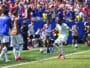 Soccer player in light kit celebrates near the touchline as teammates applaud and fans cheer in the background during a match under sunny skies.