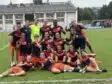 Screenshot | AllezPaillade.com Group of young female soccer players posing on a grass field in navy and orange uniforms, smiling and making victory signs after a match.
