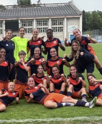 Screenshot | AllezPaillade.com Group of young female soccer players posing on a grass field in navy and orange uniforms, smiling and making victory signs after a match.