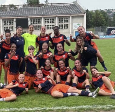 Group of young female soccer players posing on a grass field in navy and orange uniforms, smiling and making victory signs after a match.